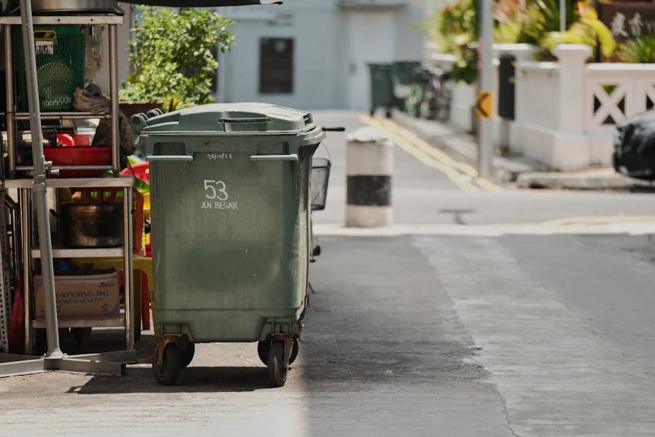 A large green wheeled waste bin with the number 53 and the words 'JUN BESAR' printed on its front, positioned on the pavement outside a property. To the left, there is a metal shelving unit holding various items including a pair of shoes, plastic containers, and a box, suggesting organisational or packing activities related to a home relocation. In the background, a street scene is visible with a road, bollards, and a white wall with decorative fencing, indicating the outside environment near a residential property. The scene is well-lit, capturing the typical process of disposing of waste during a house move or relocation, with the focus on the bin and surrounding clutter associated with moving logistics. Man with Van Mitcham often handles such tasks as part of their removals services, ensuring waste disposal aligns with local regulations during furniture transport and packing procedures.