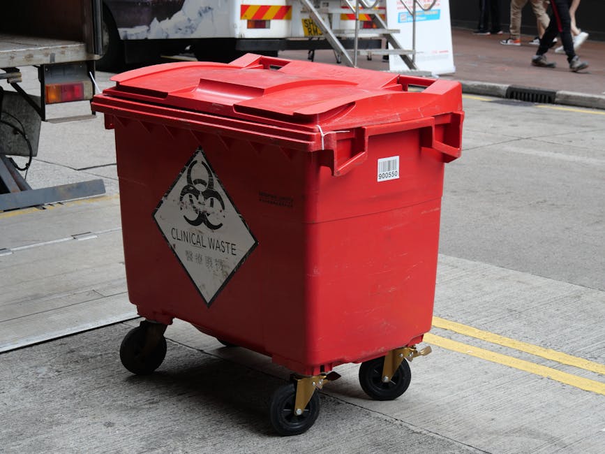 A red wheeled clinical waste bin with a black biohazard symbol and the label 'CLINICAL WASTE' in black and white, positioned on a concrete pavement during the daytime. The bin's lid is closed and secured, with visible wheels at the base enabling mobility. In the background, part of a white vehicle with a mounted stairway is visible, indicating a loading or unloading area, possibly related to house removals or relocation services. The scene appears to be outside a building or on a roadside, with pedestrians walking nearby. This image illustrates waste management procedures associated with home relocation or moving activities, highlighting the importance of proper waste disposal in compliance with local regulations, which is relevant to Mitcham's waste disposal rules for movers as discussed on manwithvanmitcham.co.uk.