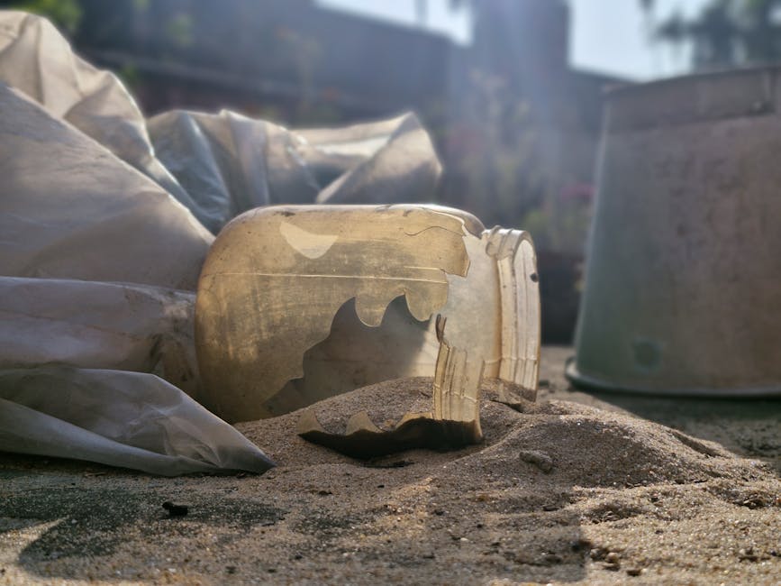 A large green wheeled waste bin with the number 53 and the words 'JUN BESAR' printed on its front, positioned on the pavement outside a property. To the left, there is a metal shelving unit holding various items including a pair of shoes, plastic containers, and a box, suggesting organisational or packing activities related to a home relocation. In the background, a street scene is visible with a road, bollards, and a white wall with decorative fencing, indicating the outside environment near a residential property. The scene is well-lit, capturing the typical process of disposing of waste during a house move or relocation, with the focus on the bin and surrounding clutter associated with moving logistics. Man with Van Mitcham often handles such tasks as part of their removals services, ensuring waste disposal aligns with local regulations during furniture transport and packing procedures.