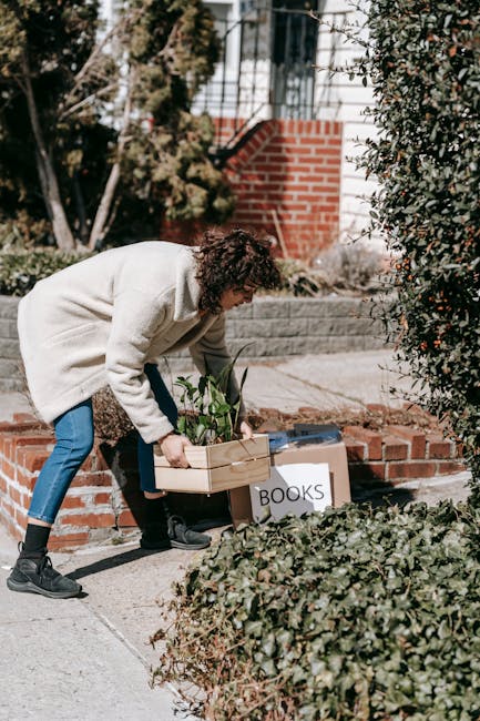 A woman with curly hair dressed in a light-colored coat, dark jeans, and black shoes is seen outdoors in a residential area during daytime, lifting a cardboard box filled with potted plants and greene