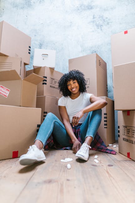 A woman with curly hair dressed in a light-colored coat, dark jeans, and black shoes is seen outdoors in a residential area during daytime, lifting a cardboard box filled with potted plants and greene