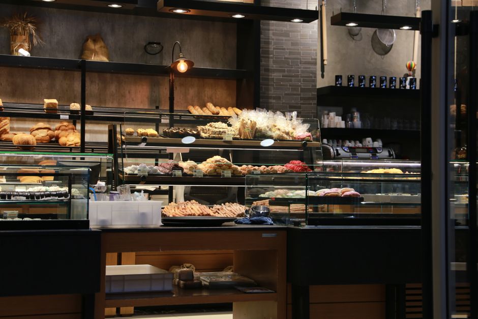 Interior view of a bakery with display cases filled with various baked goods such as bread, pastries, and cakes. The display units consist of glass-fronted refrigerated counters and open shelving, wit