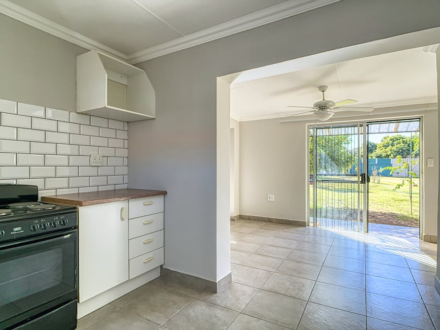 Interior view of a residential kitchen and living area during a house relocation, with a small section of the kitchen visible in the foreground showing a black freestanding oven, white cabinetry with beige handles, and a countertop with a pinkish hue. The kitchen features a white brick-style tiled backsplash and a light grey wall, with an open shelving unit mounted above. Moving into the living space, the room has large, light-colored tiled flooring and white walls, with a ceiling fan installed. To the right, a sliding glass door with vertical security bars leads to a grassy backyard with trees and a blue fence, providing natural light that illuminates the empty room. This scene captures the process of packing and preparing for furniture transport in a home relocation, with the focus on the room’s interior layout and natural light, as part of the moving logistics managed by Man with Van Mitcham.