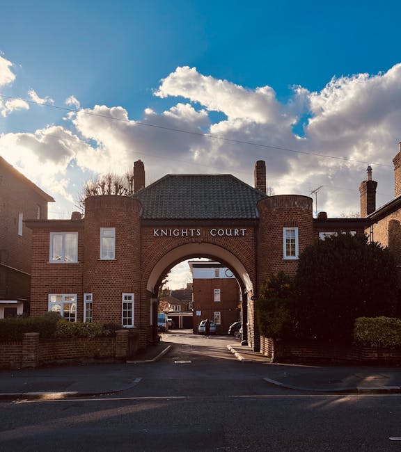 Photograph of the entrance to Knights Court, a residential property with a brick archway that has the name 'Knights Court' displayed above. The archway connects two brick buildings, and through it, a paved driveway is visible, leading to parked cars and additional residential structures in the background. The sky above is partly cloudy with bright sunlight illuminating parts of the scene. The surrounding area includes bushes and trees, and the scene appears to be taken during the day. This type of property is typical in urban or suburban areas where house removal services by Man with Van Mitcham may operate, involving the careful transportation of furniture and boxes through such pathways during a home relocation process.