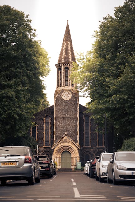 A narrow street in front of a traditional brick church with a tall, pointed steeple and a large clock face, flanked by dense green trees. Several parked cars line both sides of the street, with a visible white dashed line marking the road center. The scene is during daylight, with soft lighting and shadows cast by the trees. In the foreground, a moving van or vehicle from Man with Van Mitcham is positioned nearby, indicating ongoing home relocation or furniture transport activity, consistent with professional removals and packing services for moving from Church Road CR4. The setting suggests a residential or historic area with limited street width, requiring careful loading and logistics management.