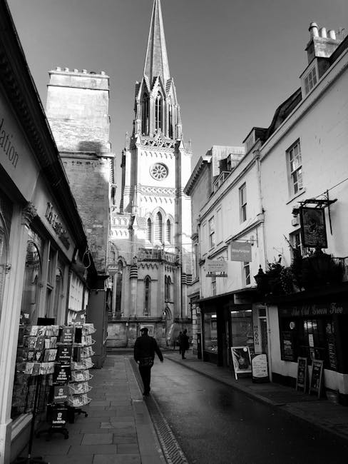 An aerial view of a residential neighbourhood with a row of closely spaced houses and narrow streets, surrounded by trees with vibrant autumn foliage in shades of orange, red, yellow, and green. The rooftops of the houses are diverse in design, with some flat and others pitched, made of materials like slate, tile, and metal. A paved street runs horizontally across the top of the image, with parked cars visible along the curb. In the foreground, a moving van marked with 'Man with Van Mitcham' is parked on the street below, with two individuals loading or unloading furniture wrapped in protective plastic and blankets into the van. Cardboard boxes and packing materials are placed on the pavement beside the van, indicating a home relocation process. The scene captures the logistics involved in furniture transport and packing within a dense, tree-lined neighbourhood, highlighting the challenges of moving on narrow streets as part of professional removals services.