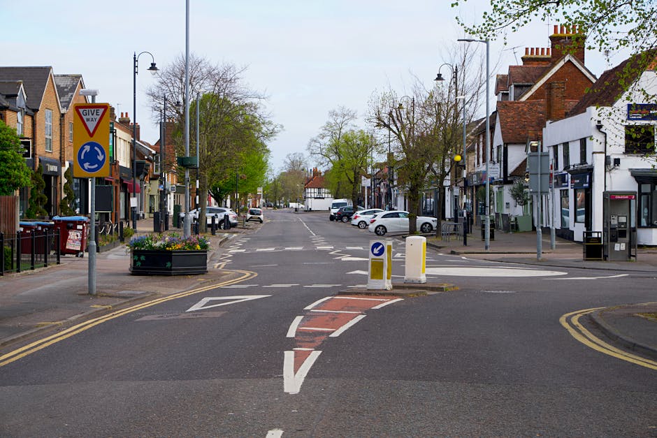 A quiet urban street scene showing a wide roadway with various parked cars along the sides, flanked by retail shops and residential buildings with brick and white facades. In the foreground, there are traffic control barriers and a red and white painted area indicating a loading zone or restricted parking, relevant to parking and loading issues on London Road, Mitcham. Street lamps, road signs including a give way sign and a roundabout sign are visible, with some greenery in trees and planters along the pavement. The atmosphere appears calm with no pedestrians or moving vehicles, highlighting the challenges of parking and loading in this area. This setting relates to house removals and furniture transport services offered by Man with Van Mitcham, with a focus on logistics and city street conditions for home relocation or moving processes.
