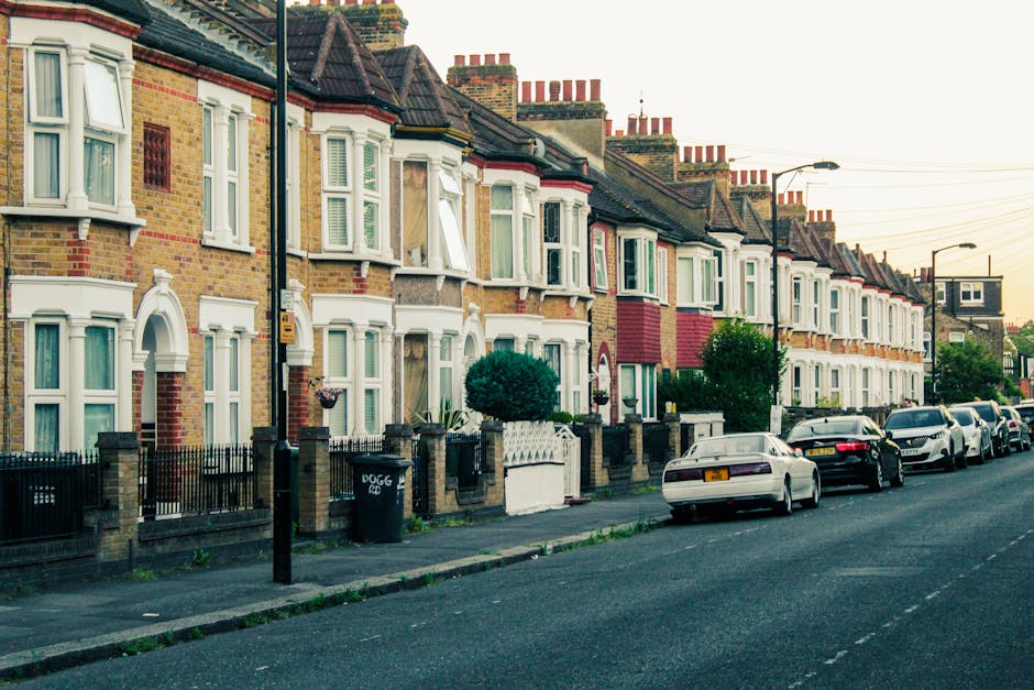 A row of terraced Victorian-style houses on London Road, Mitcham, with brick facades, bay windows, and pitched tiled roofs. The houses feature white window frames and decorative detailing, with small front gardens enclosed by black metal fences and brick pillars. Several parked cars, including a white hatchback, silver sedan, and other vehicles, line the street in front of the homes. Sidewalks run alongside the road, with street lamps and a bin visible near the curb. The environment appears calm during late afternoon or early evening, with warm lighting illuminating the scene. The image captures elements relevant to house removals and moving logistics, such as parking and access for loading or unloading furniture and boxes, supporting the context of home relocation services offered by Man with Van Mitcham.
