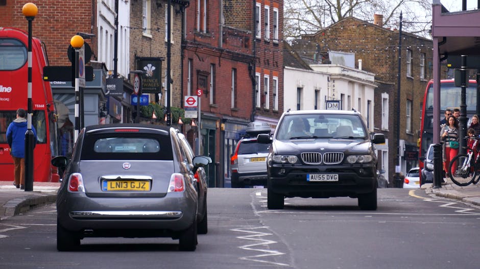 A quiet urban street scene showing a wide roadway with various parked cars along the sides, flanked by retail shops and residential buildings with brick and white facades. In the foreground, there are traffic control barriers and a red and white painted area indicating a loading zone or restricted parking, relevant to parking and loading issues on London Road, Mitcham. Street lamps, road signs including a give way sign and a roundabout sign are visible, with some greenery in trees and planters along the pavement. The atmosphere appears calm with no pedestrians or moving vehicles, highlighting the challenges of parking and loading in this area. This setting relates to house removals and furniture transport services offered by Man with Van Mitcham, with a focus on logistics and city street conditions for home relocation or moving processes.