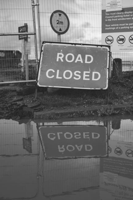 A black-and-white photograph showing a large rectangular 'Road Closed' sign positioned on a muddy ground in front of a construction site or roadworks area. Behind the sign, there is a metal fence with