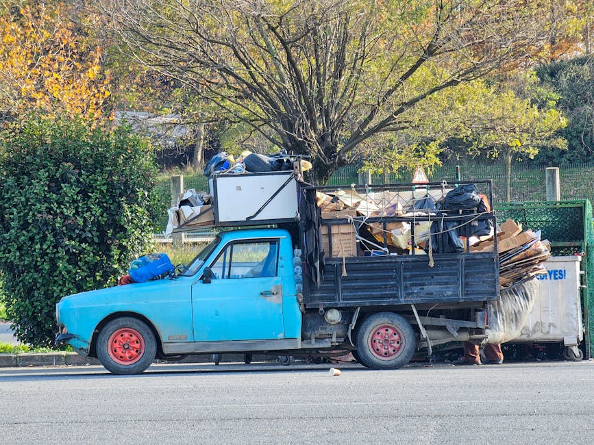 A side view of an older, compact blue flatbed truck parked on a residential street, loaded with a large amount of bulky waste including cardboard boxes, plastic bags, wooden planks, a microwave, and various household items. The truck's bed is filled with scattered items secured with ropes and plastic wrap, indicating in the process of a home relocation or clearance. Behind the truck, a green hedge and a large deciduous tree with yellowing leaves are visible, suggesting an autumn setting. The truck's red wheels contrast with the blue cab, and the vehicle appears to be part of a professional removals service such as Man with Van Mitcham. The scene captures the loading or unloading phase typical of furniture transport and packing activities involved in house removals, with equipment like trolleys possibly nearby, and the setting consistent with environmental considerations for waste disposal before moving.
